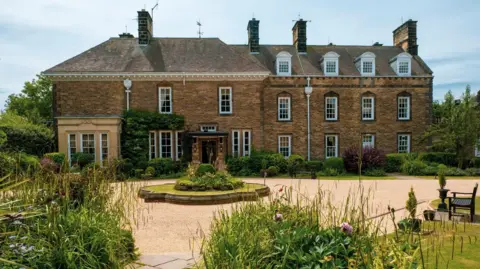 The outside of Kirklevington Hall with a wide drive and a grass planter in the middle in front of a large two storey stone building.