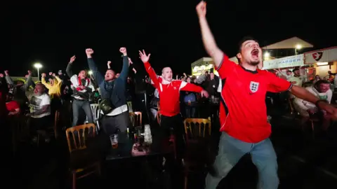 PA Media A group of male England football fans in Cheltenham jumping up in celebration while watching a football match.