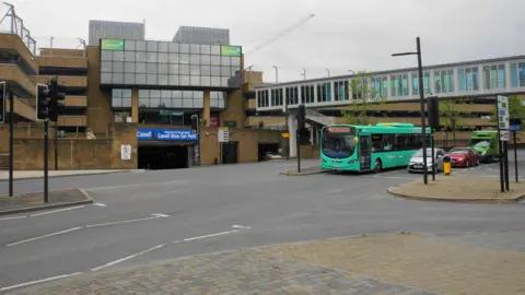 Bill Boaden/Geograph Cavell Blue Car Park, view from across the road, also shows a blue-green bus and three cars waiting in traffic, and the footbridge leading from the car park to the shops