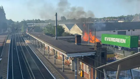 John Garfoot A fire near Stowmarket railway station. Behind the station there are orange flames at a yard with a green EVERGREEN container on a trailer parked just next to the flames.