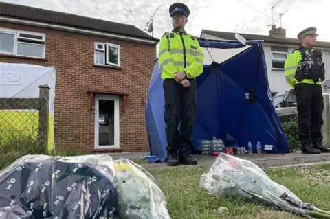 Sam Read/BBC A picture taken from the ground. In the foreground, are three or four bunches of flowers lying on the grassy front lawn. In the background, two police officers guard a bricked semi-detached house.