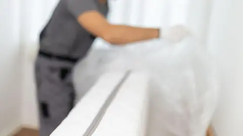 A service worker is removing the plastic covering from a mattress in a home. The room has light-coloured walls and wooden flooring. The activity takes place during the daytime. This is a stock image.