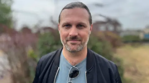 A man with long grey hair pulled back, and grey and dark facial hair. He is wearing a light blue grey t-shirt with a black zip jacket. There are aviator sunglasses hanging off the neck of the t-shirt