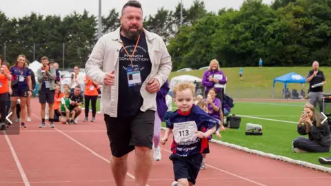 Family handout Kacey is pictured running along a sprinting track. There is a man directly behind him and other people in the background. He has a shirt with the number 113 on it. There is a woman sitting on the grass next to the track. She is taking a picture of the runners.
