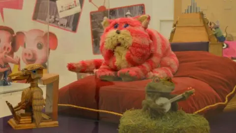 Pink and white striped cat, Bagpuss with frog, woodpecker and mice seen in the background.