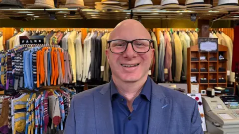 Grace McGrory / BBC Mark Edwards standing inside his menswear shop with racks of clothes behind him. He has a shaved head and wears glasses and a blue shirt and jacket