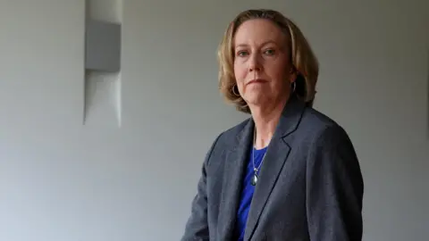 Reuters Newly appointed BP chief executive Meg O'Neill wears a grey suit and blue top while posing sat on the edge of a boardroom table.