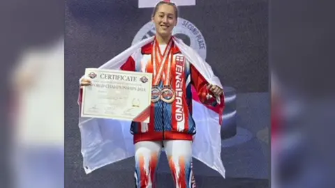 Young Somerset A girl wearing a red and blue tracksuit, draped in an England flag, holding a certificate