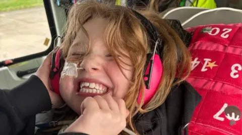 Family Amelia in a helicopter. She has a big excited smile on her face. She has a tube up her nose and large pink headphones.
