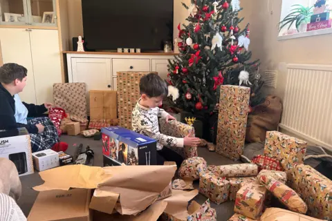 Sherianne Wood Two boys opening Christmas presents. They are both sat on the floor and there are several presents wrapped up and some opened wrapping paper on the floor as well. A Christmas tree is in the corner with red and white decorations. Also there is a white cabinet with a large TV on it. 