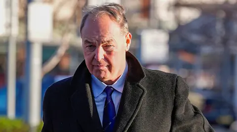 PA Media Carley, who has silver hair, walking wearing a suit, blue shirt and blue tie and a long dark coat past some railings with a bag under his left arm