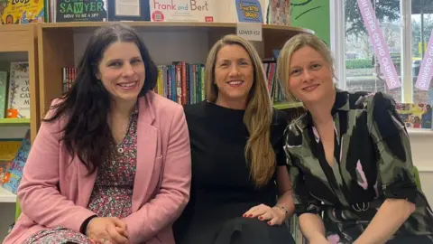Sofía Luis-Hobbs/BBC Three women look towards the camera and smile in front of a full bookshelf. The brunette woman on the left wears a floral dress under a pink blazer. The dark blond woman in the middle wears a black short sleeve dress and wears her hair down over her right shoulder. The blond woman on the right wears her hair up and wears a shirt style long sleeve dress which is a black and green abstract pattern.