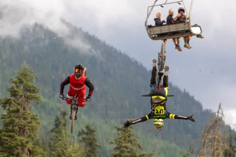 David Hanson Two bikers in the air, one upside down with his arms out. Three people on a cable car watching from behind and taking a photo. 