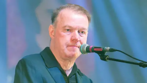 Getty Images Edwyn Collins duduk di atas panggung di mikrofon - dia mengenakan jaket biru dan t -shirt hijau dan memiliki rambut cokelat yang menipis