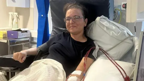 Sofia sits in a hospital chair. She has glasses and is smiling, with her hair tied back. Blood is being taken from her left arm.