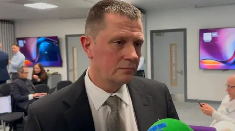 Det Ch Insp Pye, with short mousey coloured hair and wearing a dark grey suit, speaks to reporters in a busy briefing room