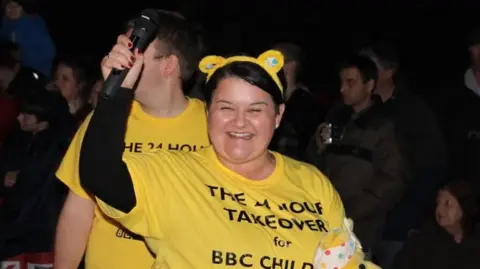 Emma Britton smiles as she raises a microphone in the air. She is wearing a yellow Children in Need top.