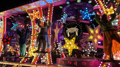 A cart with people standing on it in costume. The cart is covered in brightly coloured shapes and lights.
