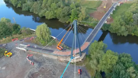 An aerial view of a bridge, midway through construction, over a river with trees on both banks and construction vehicles in the foreground.