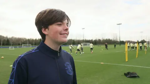 Mackenzie, who has brown hair and wears a zip up Everton branded jacket, smiles at the camera as Everton players train on a grass pitch behind him