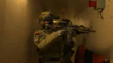 Ministry of Defence A screengrab from a video which depicts an armed forces personnel in tactical gear holding a gun. They are in a small corridor and wearing a helmet and gloves with most of their face covered.