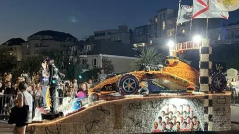 A large model of a Formula 1 car made of yellow flowers. It is lit up and there are spectators around it.