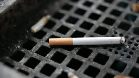 BBC Close-up of smoking cigarette on the square grill of a public ashtray.