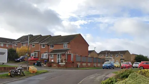 Mill Green Road on an overcast day. The houses are made with red bricks and there are cars parked along the road.