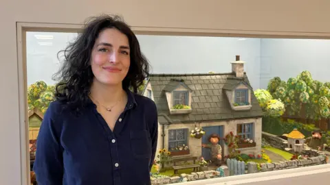 Rosy Whittemore smiles as seh stands in front of a wall dispaly showing the set of a quaint cottage from a Postman Pat sequel