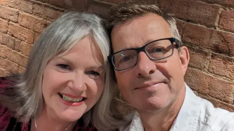Marie and Simon Haythornthwaite pose close together. She has grey, shoulder-length hair with a middle parting and wears a silver necklace. He has grey-brown hair and glasses and wears a white shirt.
