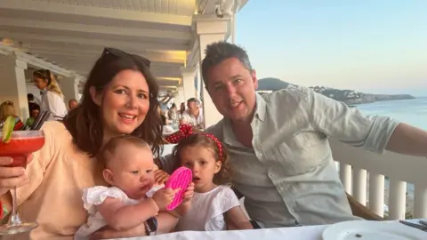 Submitted Lucy, Sienna, Margo and Tony, on holiday, with Lucy holding a cocktail, Sienna playing with a pink toy, Margo with a red hairband and Tony wearing a light coloured shirt.
