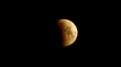 Rockboffins A blacked out image with just the Moon in vision. It is partially covered by shadow and an orange/yellow colour with some blotches of slightly darker colour.