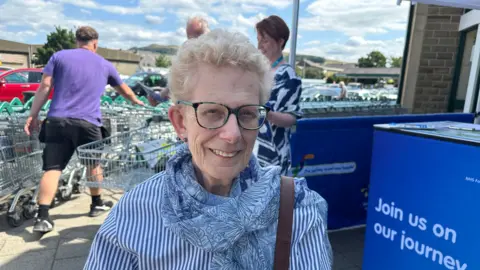 Jessica Bayley/ BBC A woman with short white hair and glasses smiling looking towards the camera. She is wearing a blue and white stripe shirt with a light white and blue patterned scarf on. Behind her are a couple of people and rows of shopping trolleys.