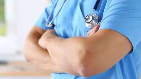 Getty Images A hospital worker, wearing blue scrubs, with their arms folded. They have a stethoscope around their neck. 