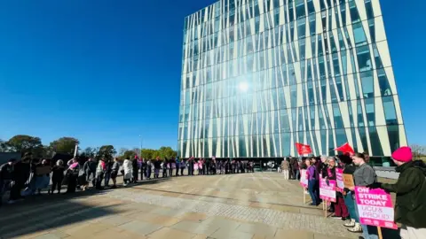 Strike action in Aberdeen outside a glass university library building, with sunshine reflecting off the windows, under a blue sky, protestors are holding protest signs.