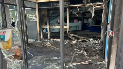 A window of a leisure centre smashed through. There is smashed glasses and burnt material lying on the floor of the reception area.