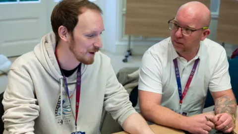 Two men sitting at a desk together, both wearing a pink, purple and blue lanyard with ID cards. One man is bearded and wearing a cream hoodie, he's smiling whilst speaking. The other man is bald, wearing glasses, and appears interested in what the other man is saying. He's wearing a white polo t-shirt and has a tattoo covering the lower part of his arm. 