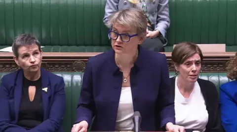 UK Parliament A woman with short light brown hair, blue glasses, a white top and blue jacked standing up in a room with dark green upholstered benches, with two women with dark brown hair and blue tops on either side of her