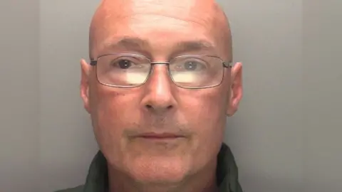 Police mugshot of a bespectacled Eoin O'Toole, who is bald and is wearing a dark-coloured top.