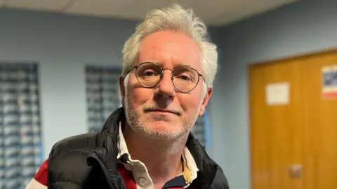 Rob Francis is wearing a red, white and blue rugby shirt and a black gilet. He is wearing glasses and has white, short hair and a short, white beard.