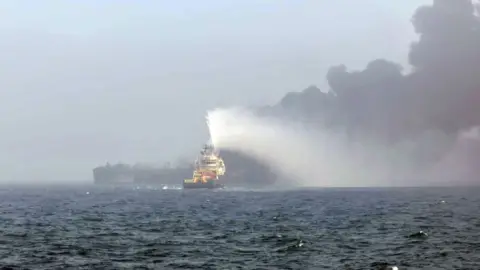 A wide shot showing a rescue boat in the North Sea heading towards the site of a oil tanker collision with a large plume of black smoke overhead.