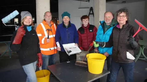 Jason Bryant Six people in a row, holding paint rollers, hammers and brushes. Either end are women, and in the middle is men. They are in an industrial-looking building.