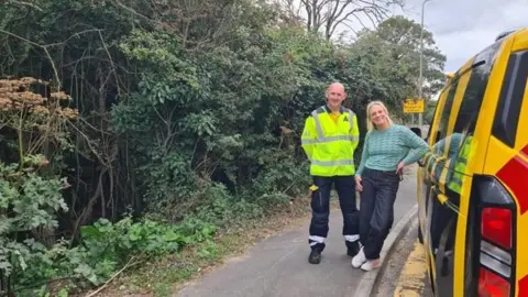 A man with a bald head and wearing a yellow high-vis jacket stands looking in to the camera. Beside him is a woman with blonde hair, a light green long-sleeved top, dark trousers and white trainers. She is shorter than him and leans again a yellow AA van. The are standing on a pavement which has thick trees and undergrowth next to it.