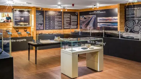 Historic England Archive A view of displays at Flag Fen Archaeological Park. It shows a pine clad interior covered in black displays boards with white writing and photographs on the walls. Against the walls are black display cabinets with artefacts within. Against the wall on the right is a long preserved log boat. In the middle of the room are two more display cabinets with artefacts under glass and replica tools. 