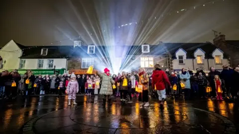 Paul Campbell A crowd of people gathers in an open space in Dingwall. It is night-time and cold, and everyone is wearing outdoor winter clothing. There is a row of traditional-style buildings behind the group and beams of light shine in the centre of the image.