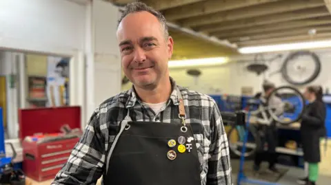 A man working on a bicycle in a repair shop. He is wearing a black apron over a black and white check shirt