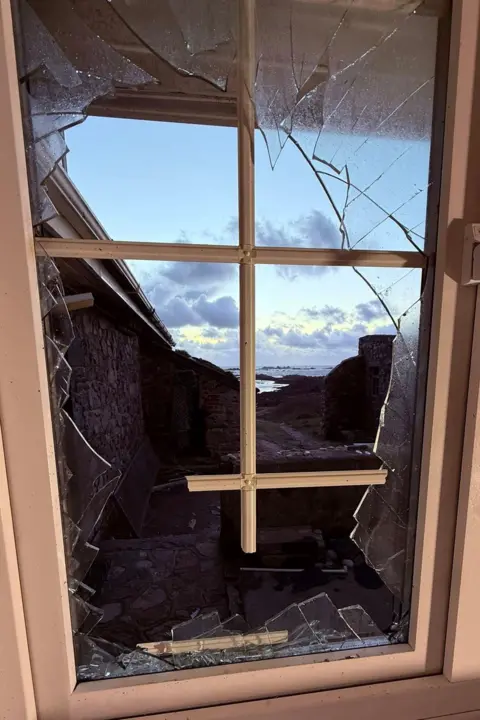 Lihou Charity LBG A window frame is left splintered with smashed and missing glass. The view looks to the sea.