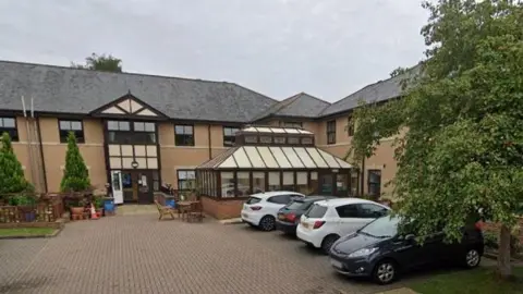 The former Hallgarth Care Home is a two-storey beige brick building with a small outside seating area and a four parked cars.