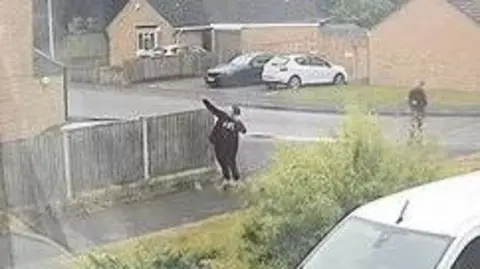 A young teenager dressed in all black with his arms outstretched, aiming a slingshot at a pigeon out of frame