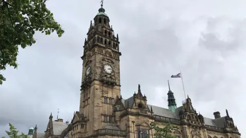 A gothic building with a clocktower, against a grey cloudy sky. Leaves from a tree in the top left of the frame.